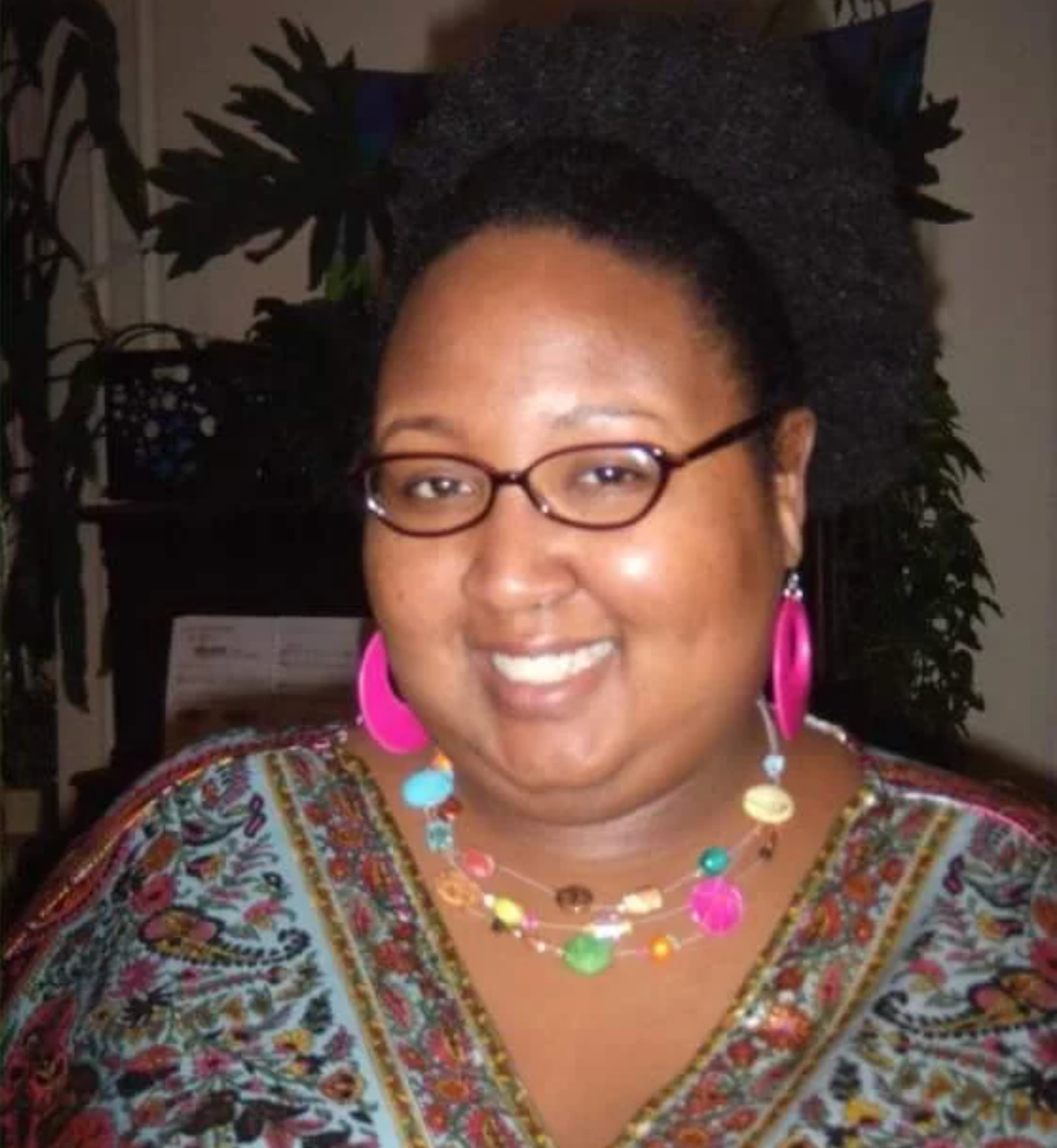 A Black woman smiling at the camera. She wears a v-neck blouse with a multicolored print and brightly colorful earrings and necklace. 