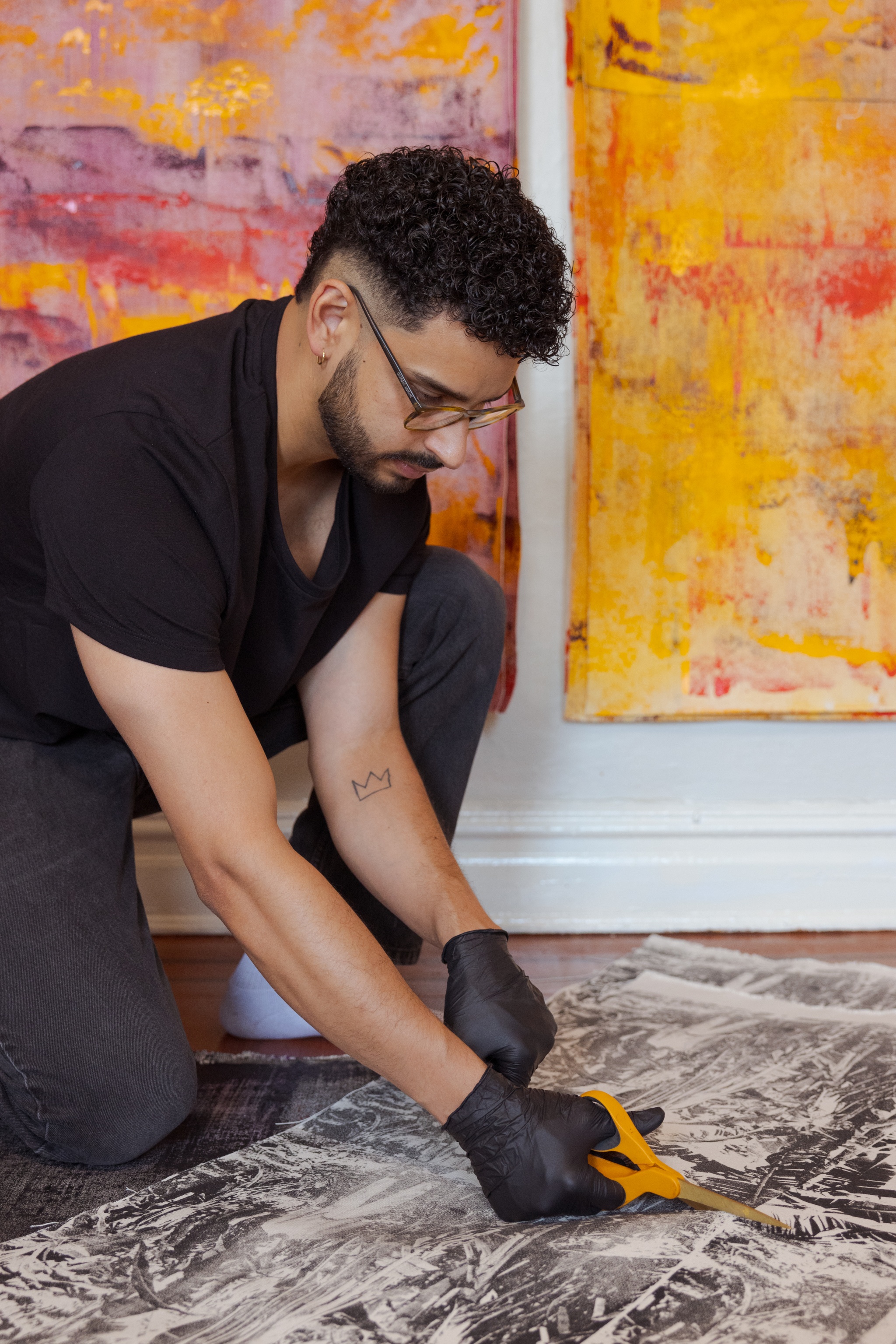 The artist Luis A. Gutierrez is seen crouching over printed canvas fabric laid on the floor. He is cutting the fabric down the middle with orange scissors.