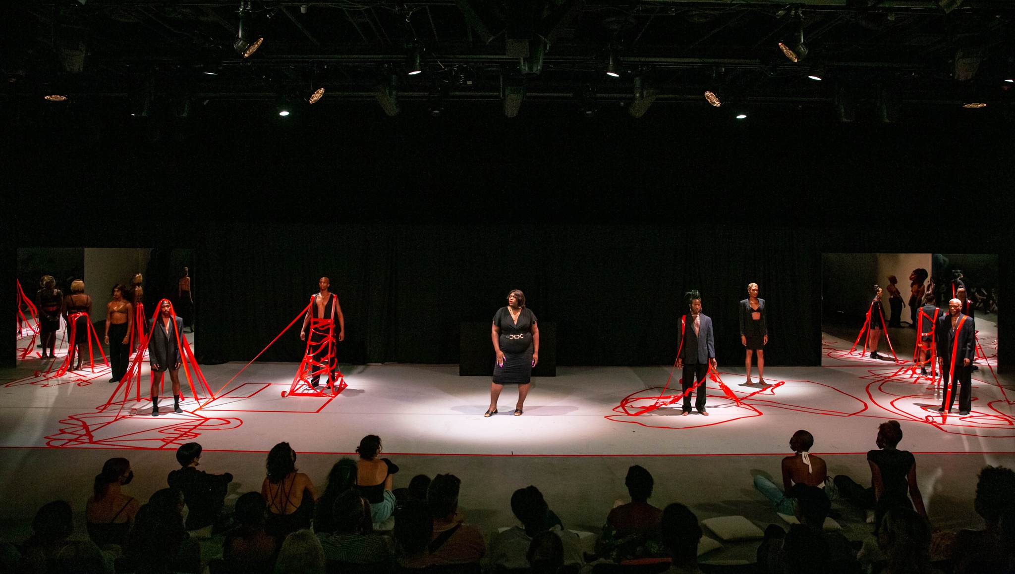 Nine Black trans performers stand on a stage wearing black suits and dresses in front of an audience. On the floor of the stage, two performers embrace as they roll together across the floor. Six of the standing performers have red tape reaching up from the ground to hold them in place.