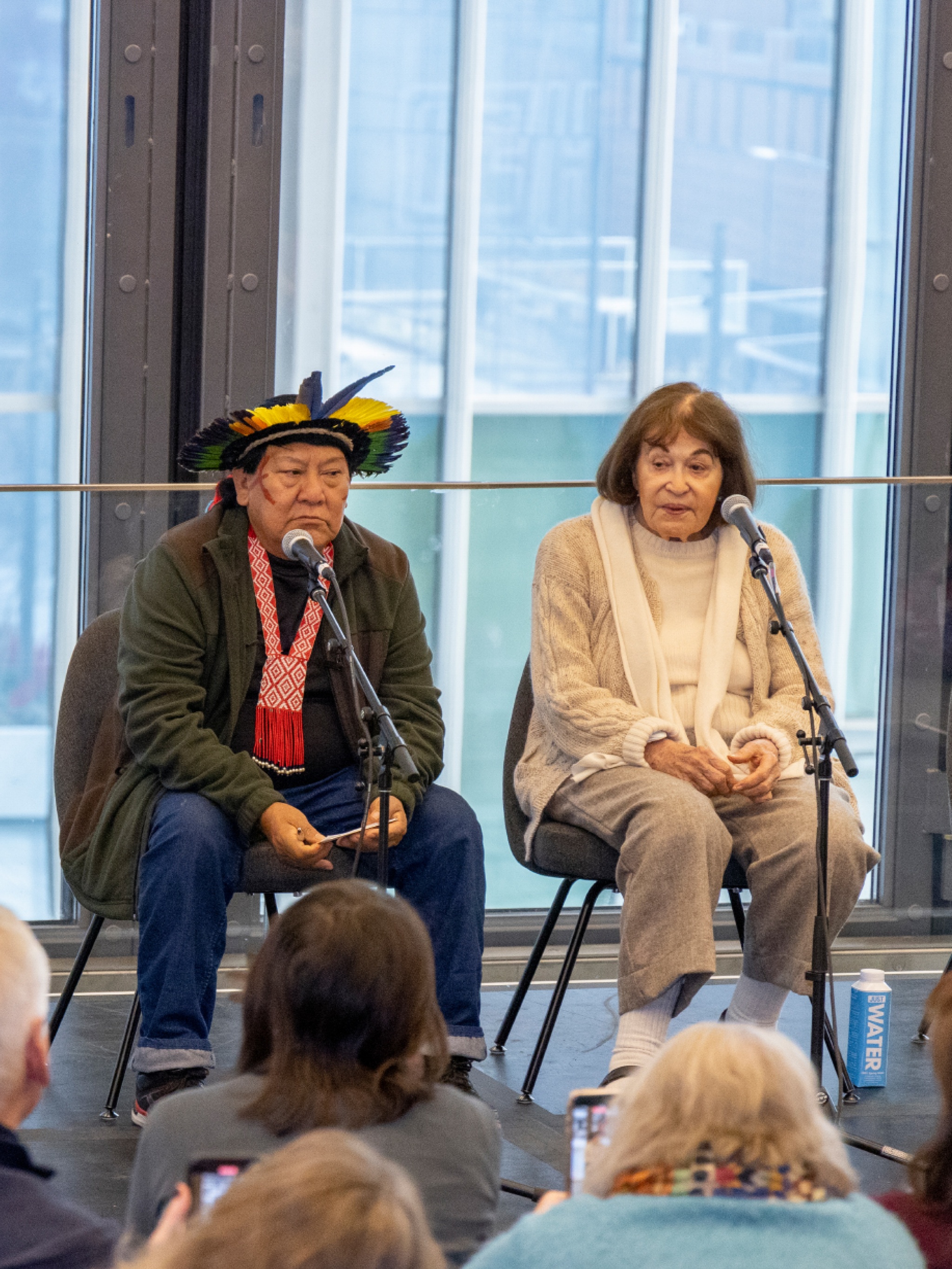 Two panelists sit on a stage with large windows behind them. On the left, is Davi Kopenawa, a Yanomami man who wears a head dress made of colorful feathers. On the right of him sits artist Claudia Andujar, a Brazilian white woman with brown hair wearing an off white outfit.