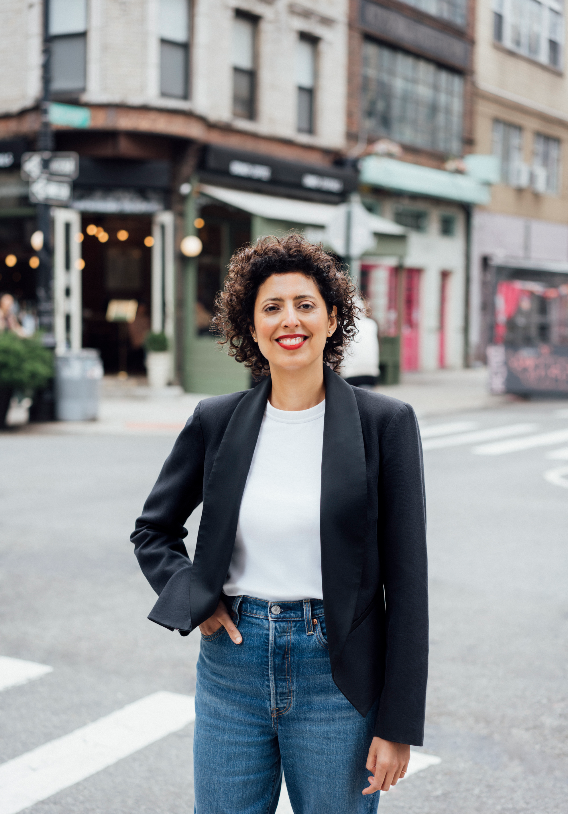 A portrait of Sara Raza standing in a city intersection with one hand in her jeans pocket. She has short curly hair and smiles at us. Photo by Asya Gorovits
