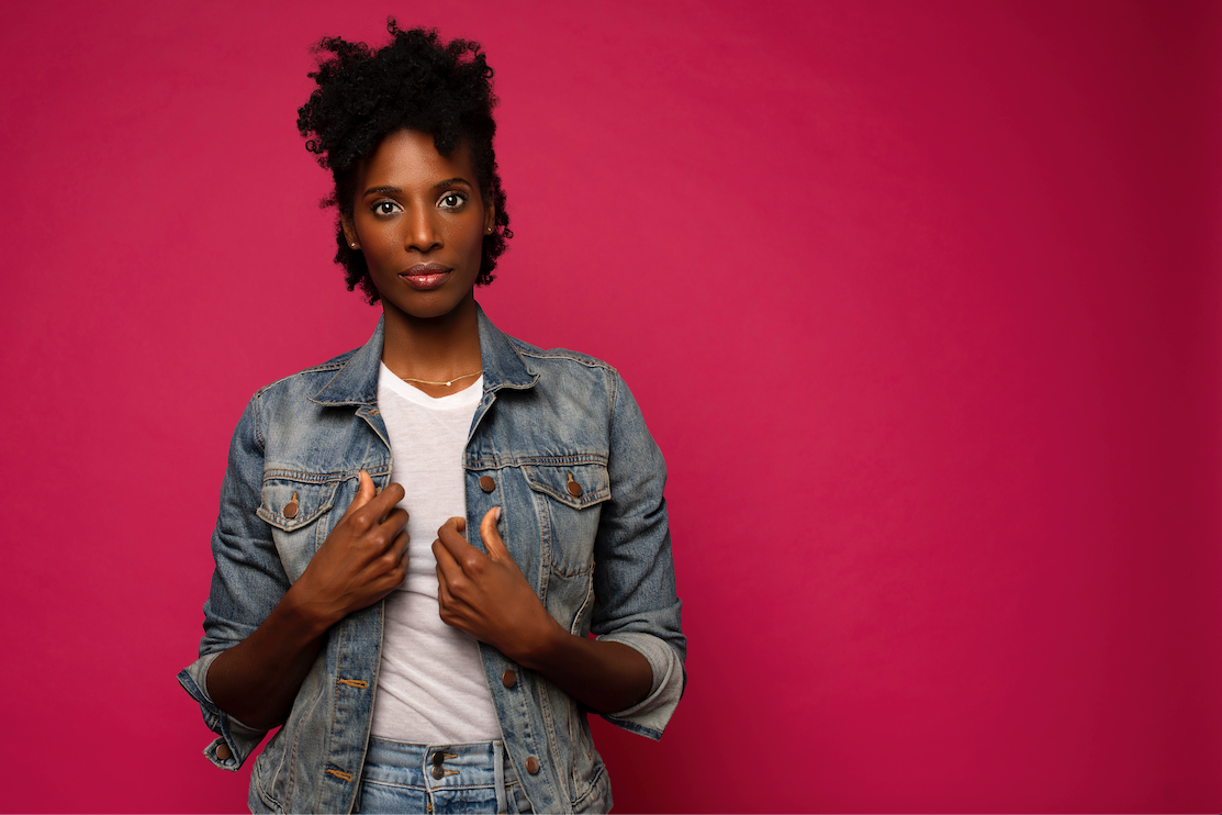 Zainab Johnson, a Black woman, standing against a bold magenta wall with her hands holding the lapels of a blue jean jacket she wears over a white t-shirt. 