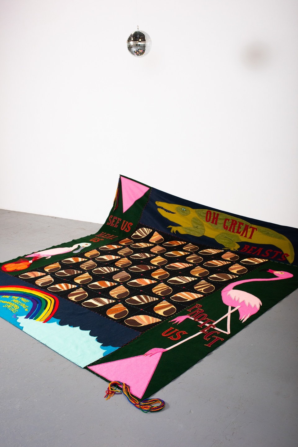 A colorful quilt is slumped up against the white wall of a gallery space. The quilt is decorated with an alligator, a flamingo, a pelican, water, a bag of oranges, two large pink triangles, a rainbow, and a grid of spiral seashells. Letters on the quilt read "Oh great beasts, see us, heal us, protect us." A miniature disco ball hangs above it.