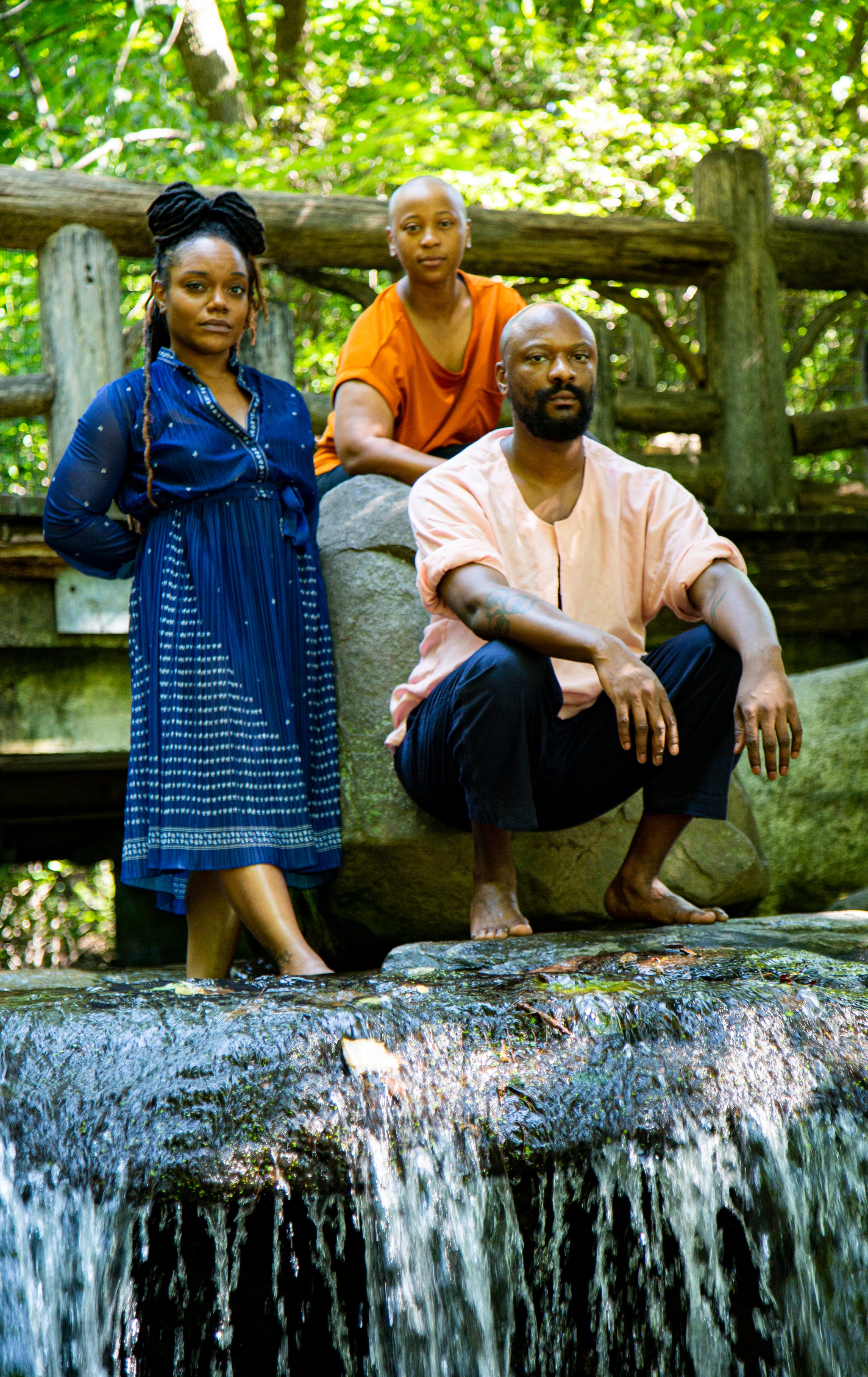 A portrait of the performance trio the HawtPlates with three people standing and crouching barefoot at the top of a small waterfall with a bridge behind them