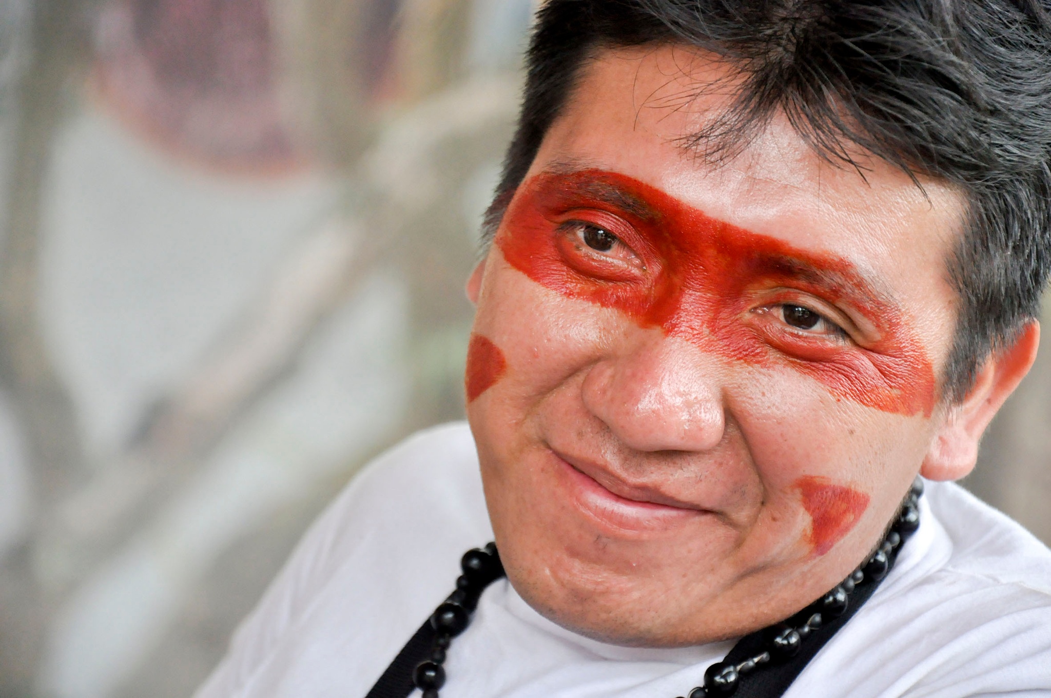 A portrait of artist Morzaniel Iramari. An Indigenous Yanomami man, Morzaniel is seen in a close-up of his face. He has short, dark hair and smiles broadly without opening his mouth and has a band of red paint across his eyes and accenting his cheeks. 
