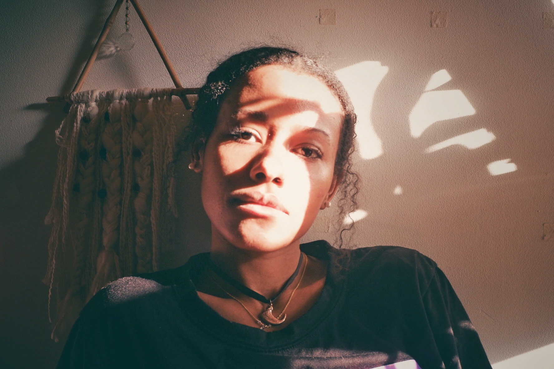 A woman sits with her face in the play of light and shadow from a window to her left. She wears a choker, as a twist of hair that falls down the left side of her face, and sits ini front of a geometric wall hanging. 