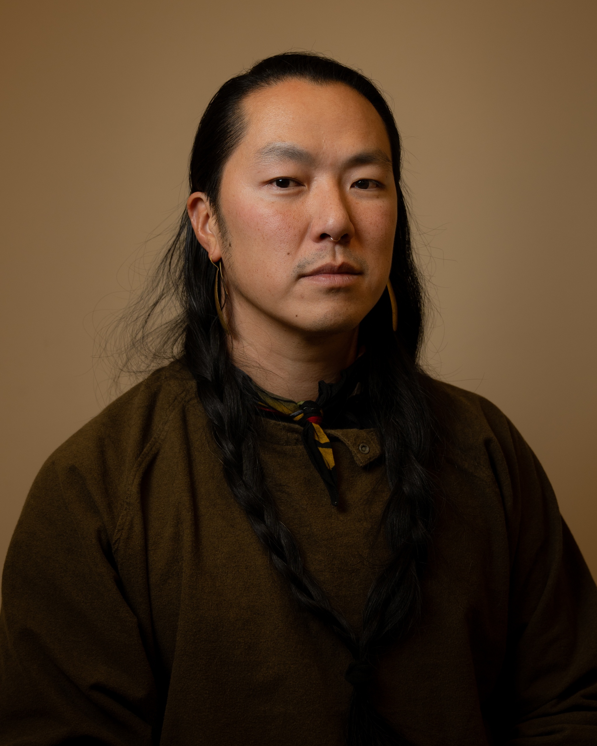 A portrait of enrico dau yang wey who poses against a neutral brown background wearing a brown shirt and jacket. enrico has long dark hair pulled back behind his head. He looks directly at us.