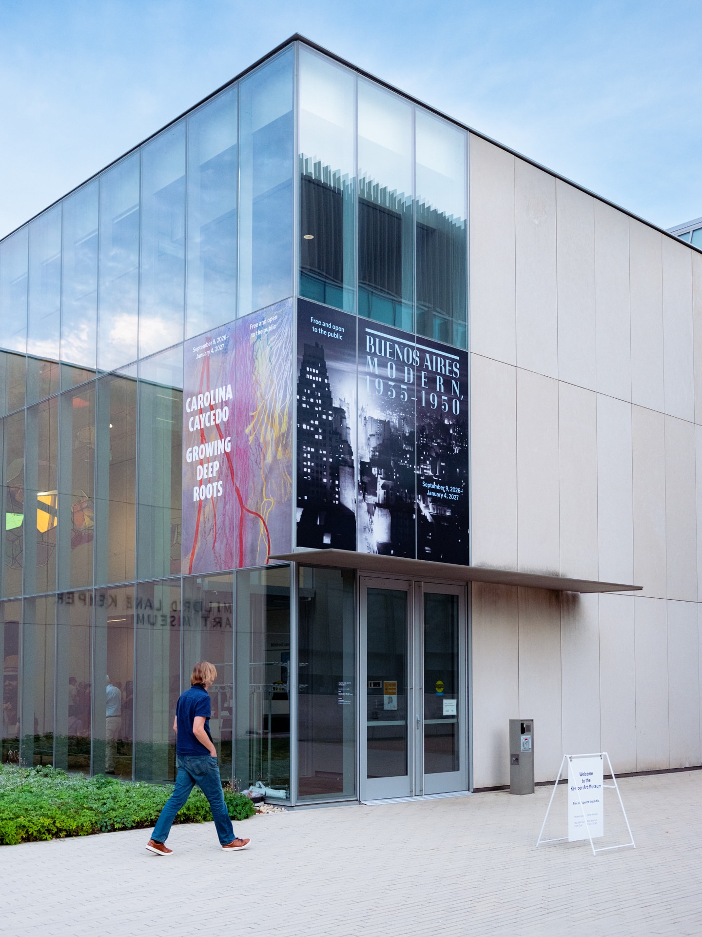 Exterior image of a man walking towards the Kemper Art Museum's entrance, which features a corner of floor-to-ceiling glass windows featuring exhibition banners.