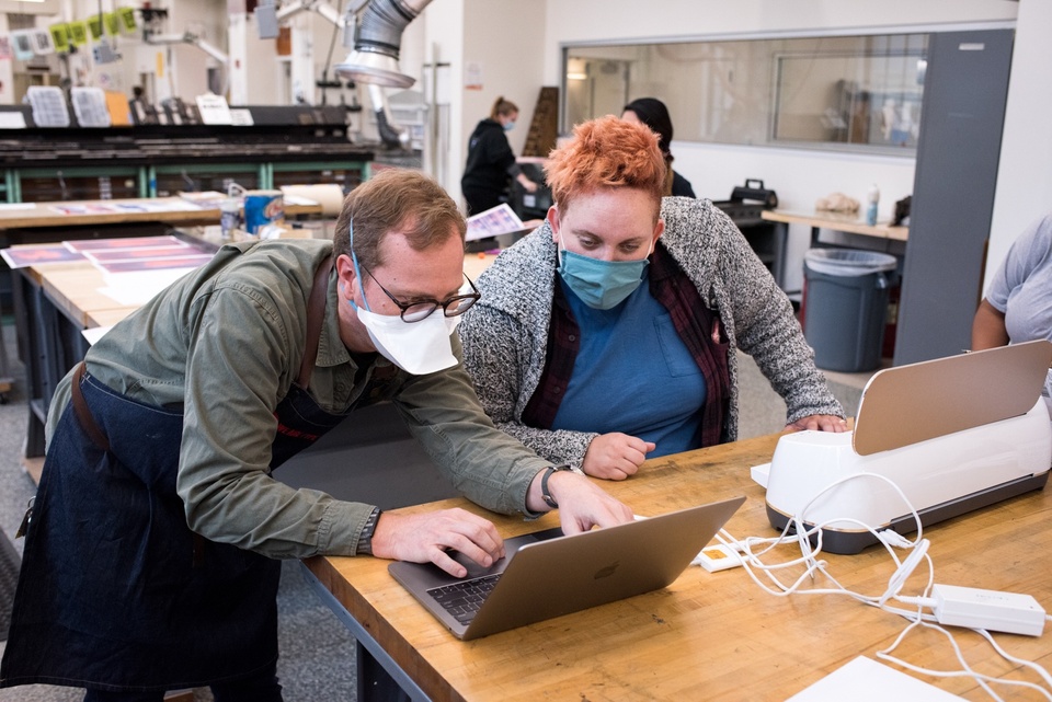 Two people look at a laptop on a worktable next to a Cricut machine.