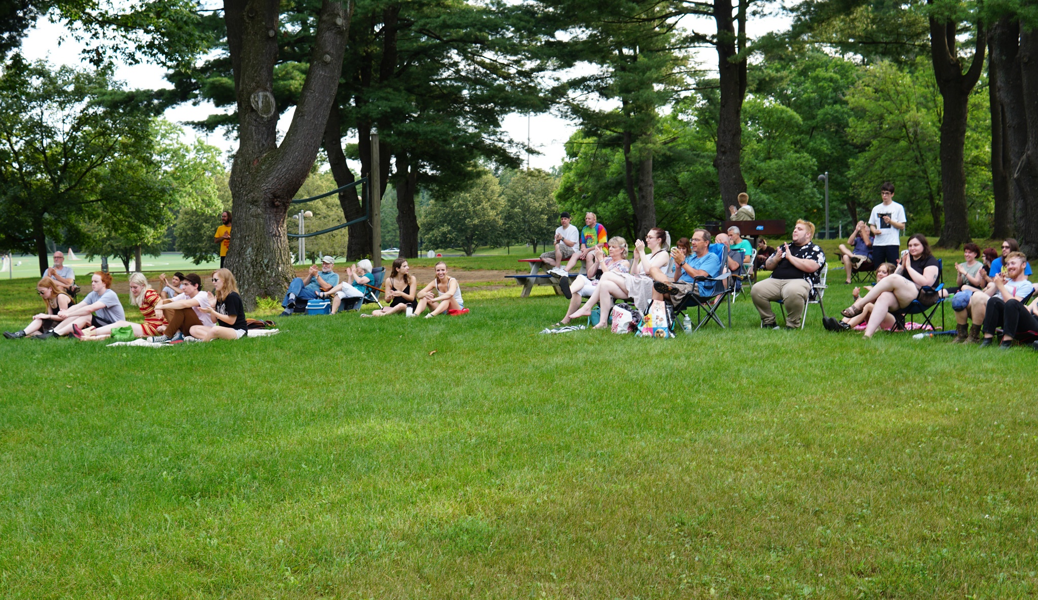 A crowd of people sit on a lawn with trees behind them, some on lawn chairs, others on blankets.