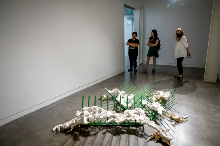 Amy Hauft and two students examine a sculpture on the ground. The sculpture is made of green crisscross shapes in metal and irregular clay feature 