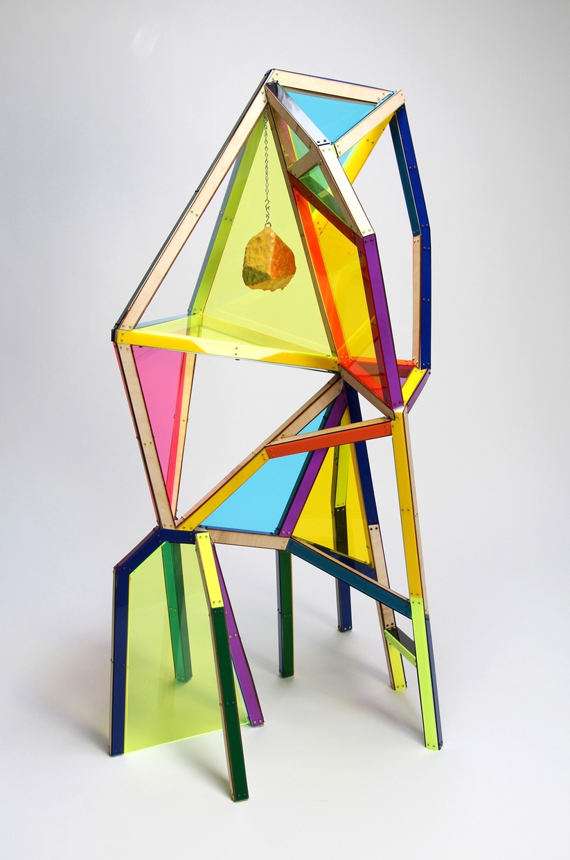 A small rock hangs from a chain inside a larger geometric structure with colorful transparent acrylic panels enclosing some areas.