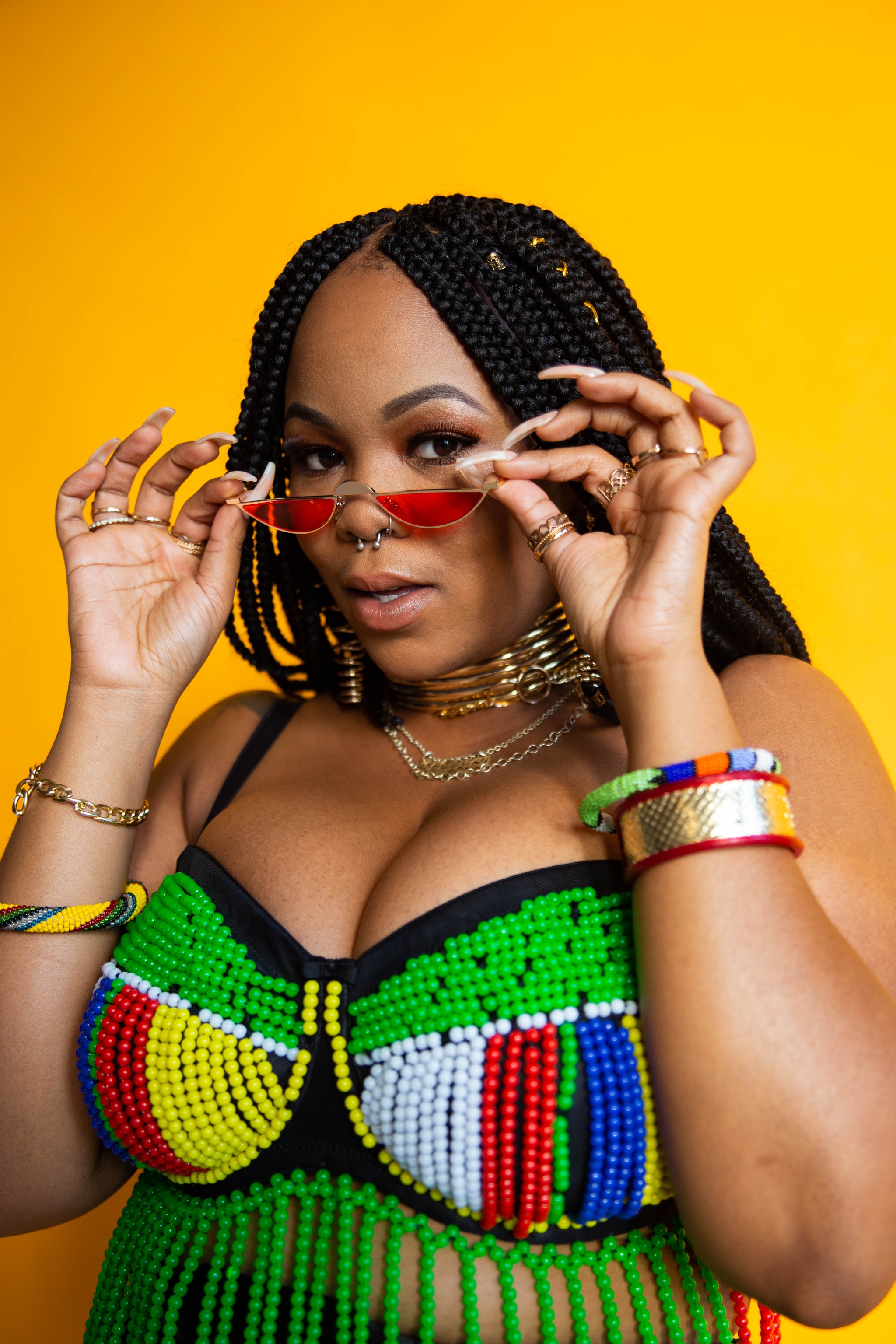 A Black woman looks out at us over a pair of red-tinted sunglasses that she pulls down with both hands. She wears colorful bracelets on both wrists and a low-cut top with green, yellow, red, and white beading.