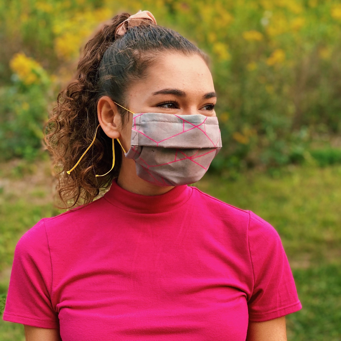 A tanned, young woman with dark hair wears a bright pink shirt and a colorful mask.