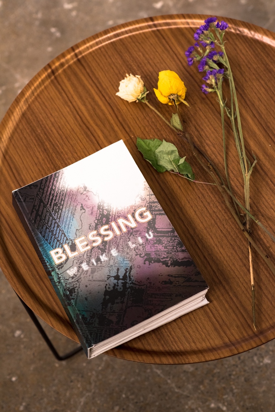 Overhead shot of a thick book on an end table decorated with dried flowers. The cover is an abstact drawing in black, blue, and purple and is titled "Blessing."