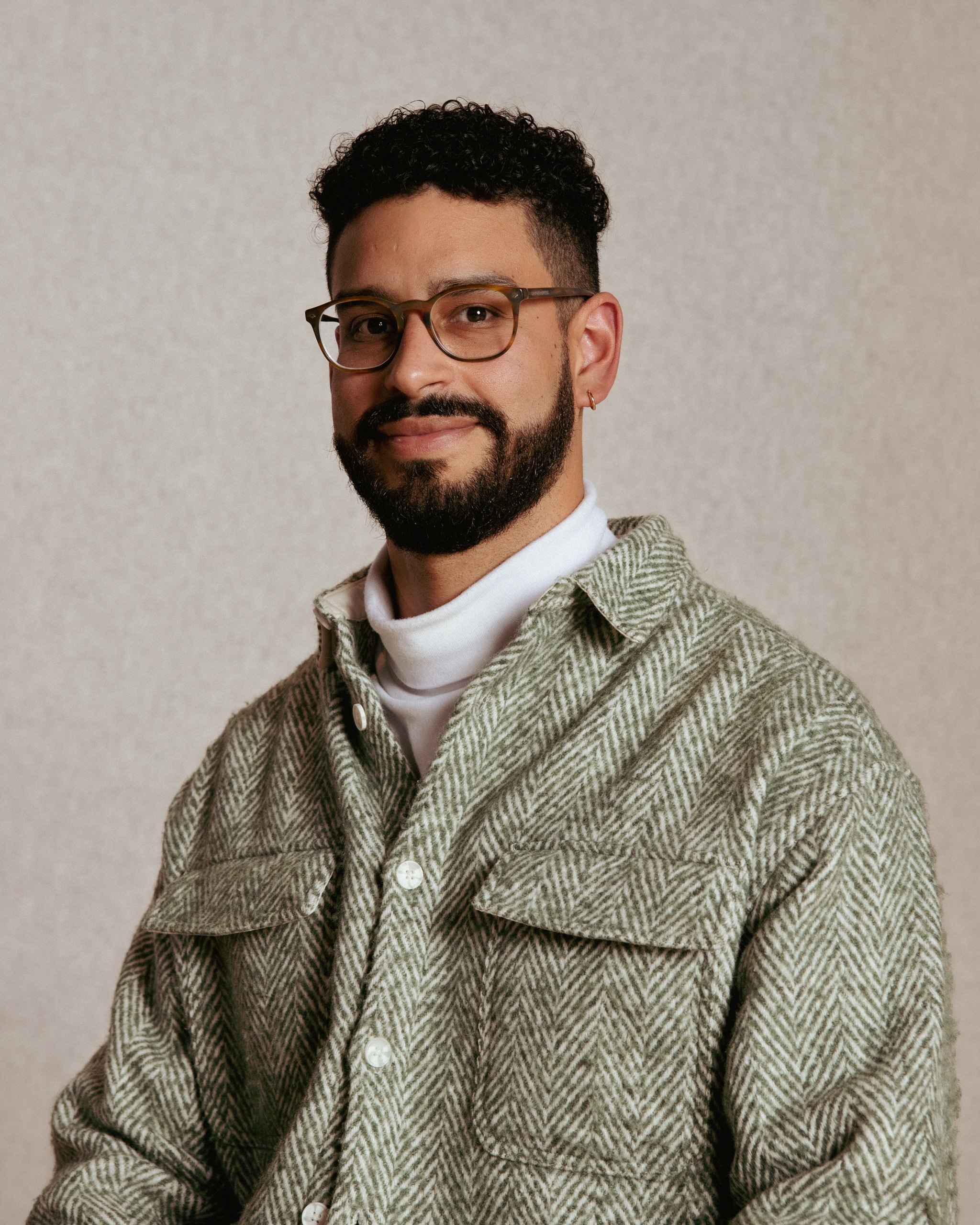 A portrait of Luis A. Gutierrez, a Colombian man who poses against a plain beige background. He sits with arms at either side, wearing a green herringbone shirt buttoned over a white turtle neck. He has a dark beard and mustache and wears glasses. Photo by Dana Golan