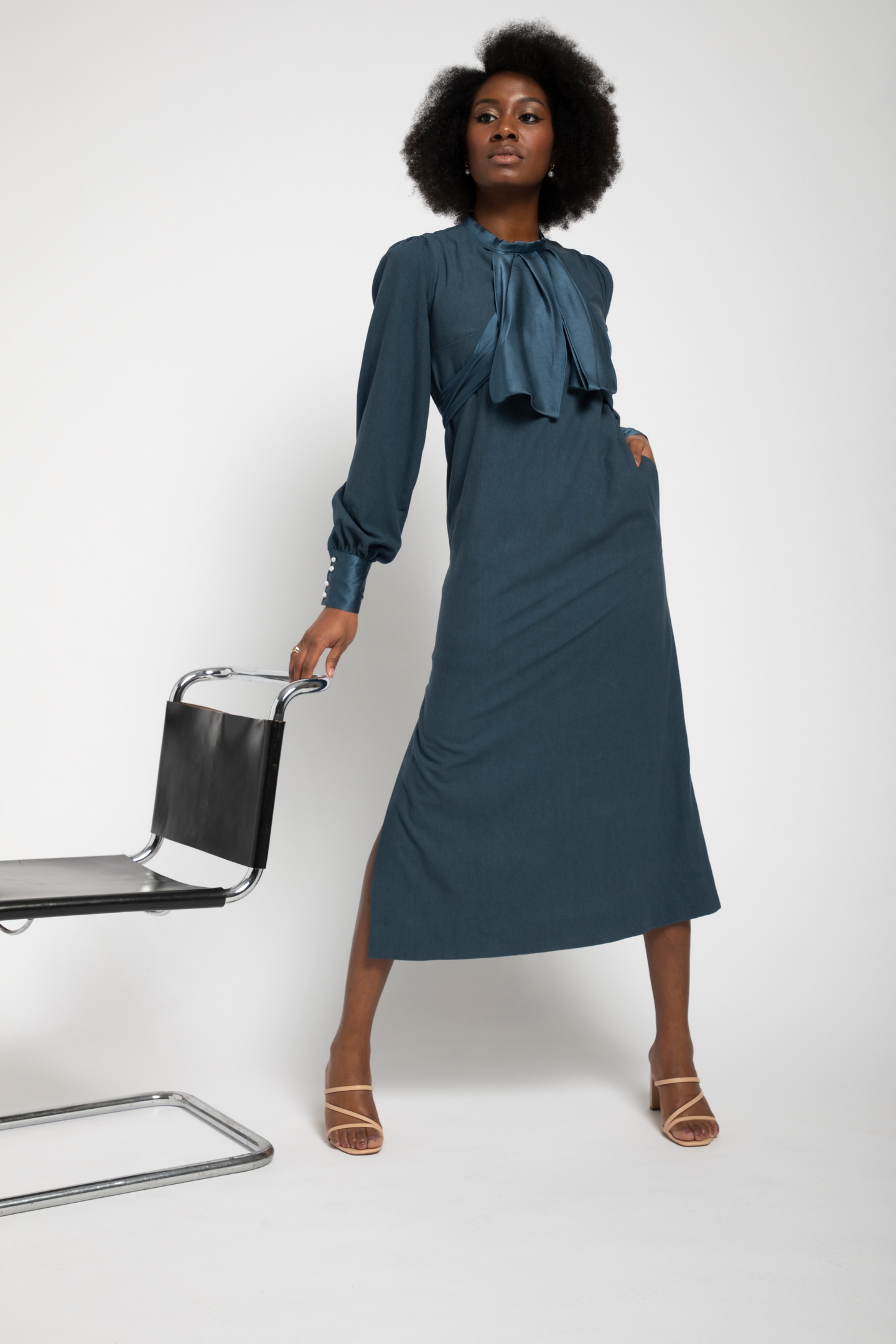 Model in a dark teal dress with long sleeves with a narrow satin cuff and a satin cravat collar. Model poses with a midcentury modern conference room chair.