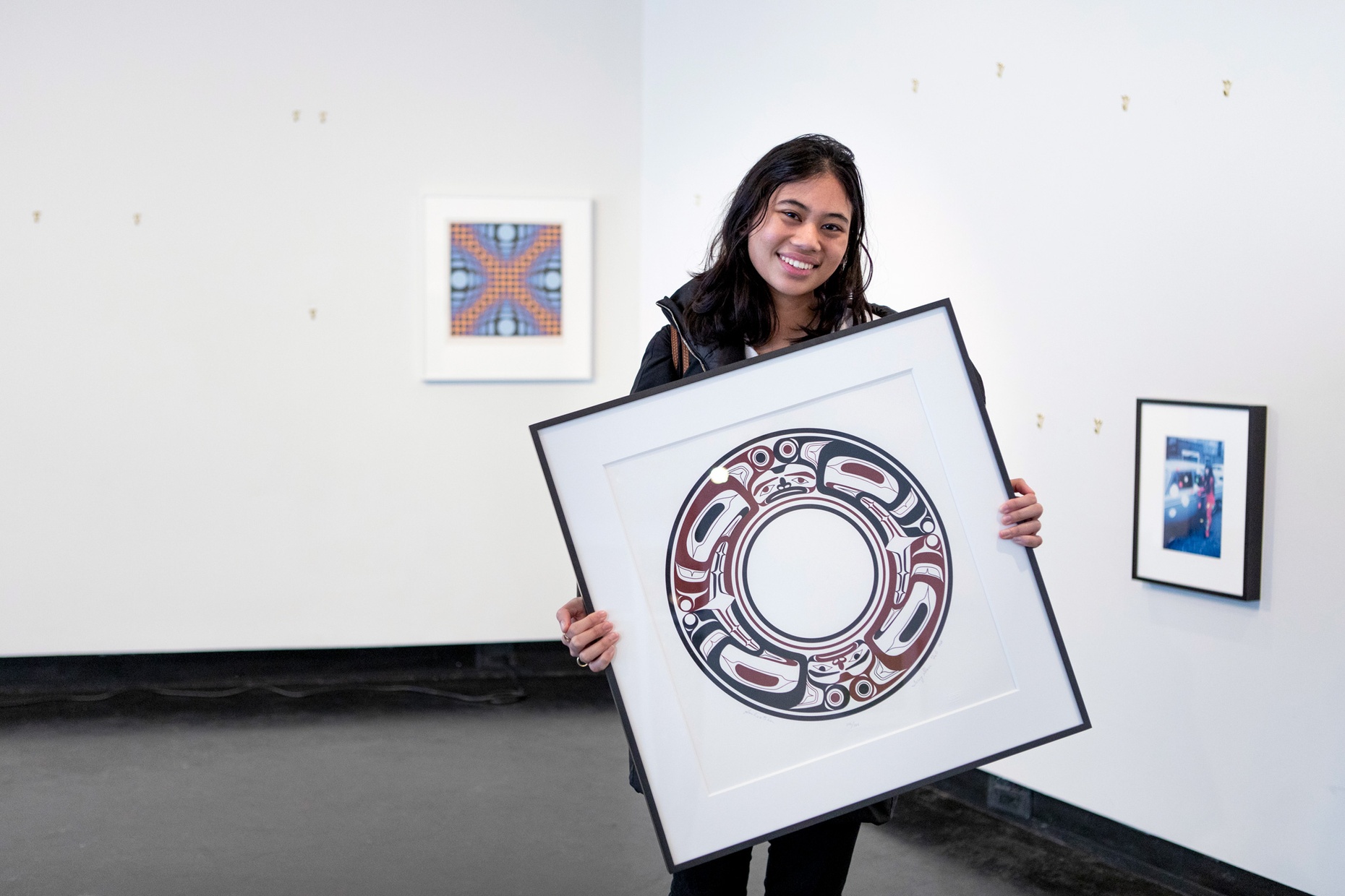 A smiling student holds a framed artwork by artist Terry Starr in her hands with two artworks hanging on the wall behind her.