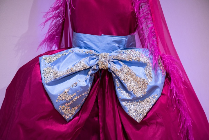 Detail of a hoop-skirted dress made from magenta satin and cinched with a huge baby-blue bow bedazzled with rhinestones and draped with a magenta veil edged in feathers.