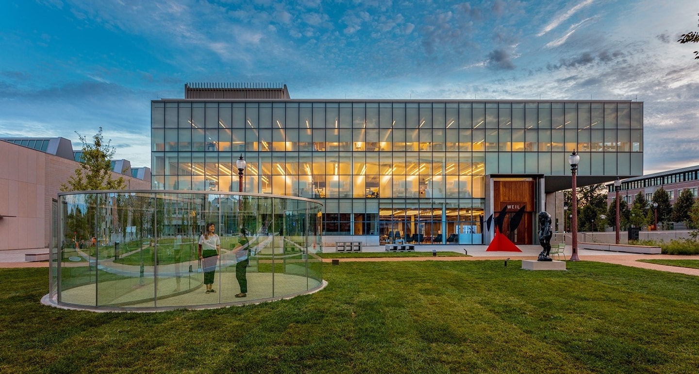 Anabeth and John Weil Hall at Washington University in St. Louis, featuring its prominent glass-curtain wall facade and the surrounding sculpture garden at dusk.