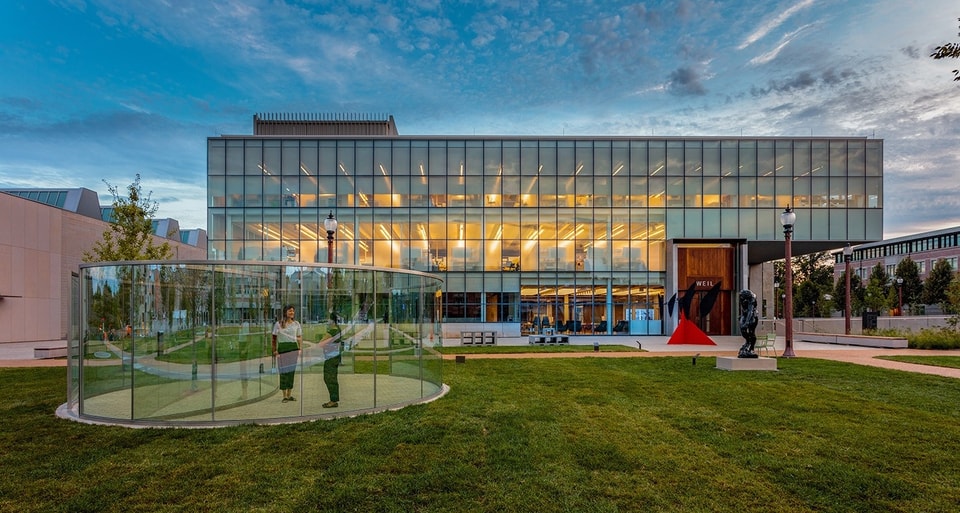 Anabeth and John Weil Hall at Washington University in St. Louis, featuring its prominent glass-curtain wall facade and the surrounding sculpture garden at dusk.