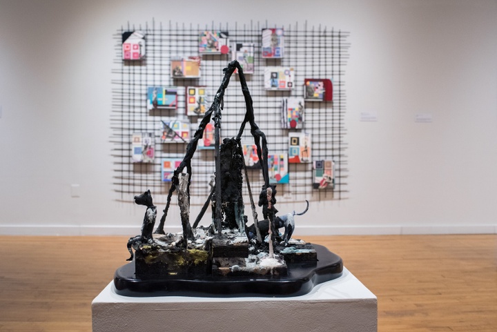 Black viney sculpture that forms a mountain-like peak sits on a white plinth. On the wall behind is a trellis with a dozen small colorful paintings affixed to it. 