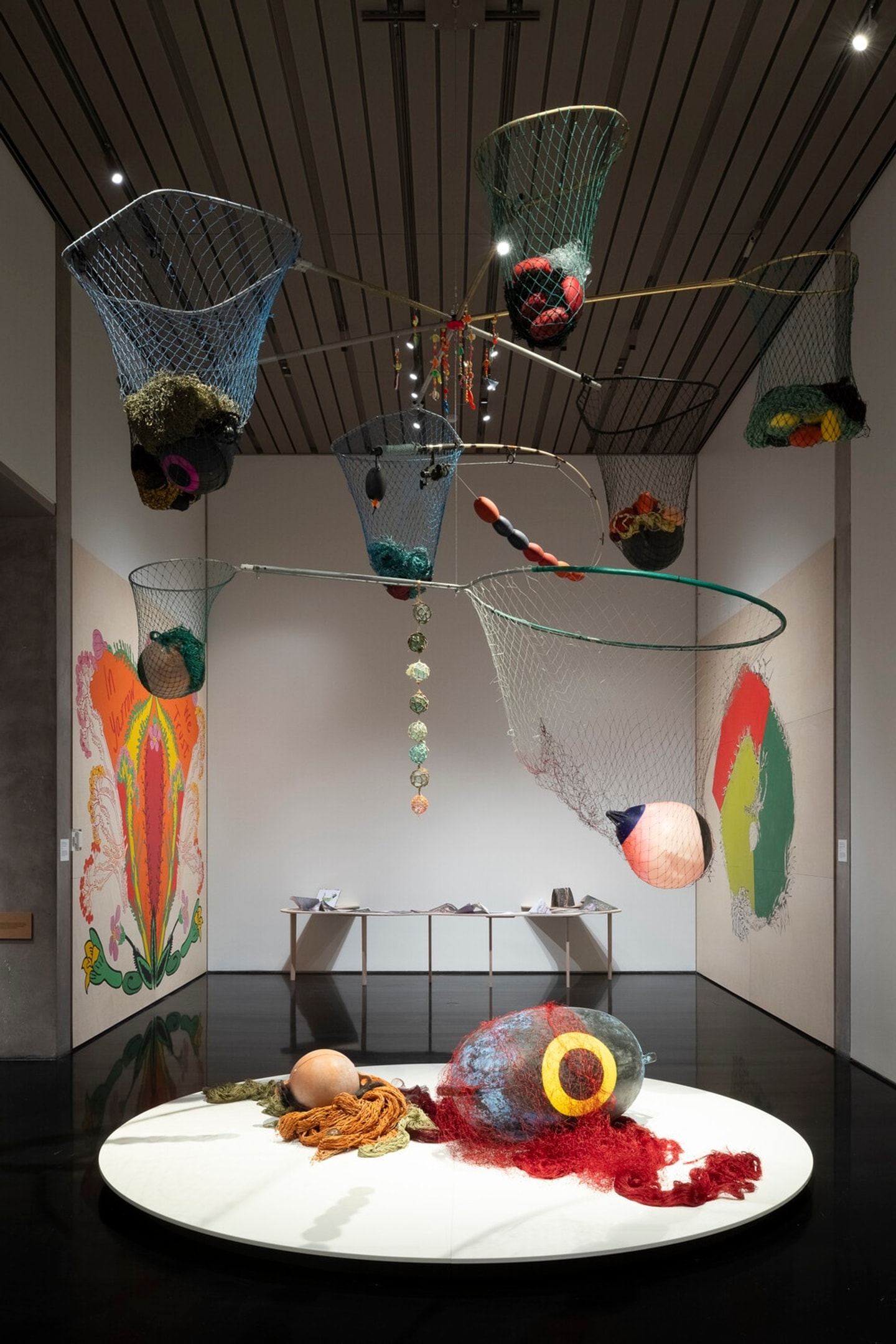 An installation view of a gallery featuring suspended fishing nets and dipnets filled with colorful buoys, fishing lures, rope, and other materials. A large netted form rests on a circular platform in the foreground, while colorful botanical prints and drawings line the walls and additional objects are displayed on a table in the background.