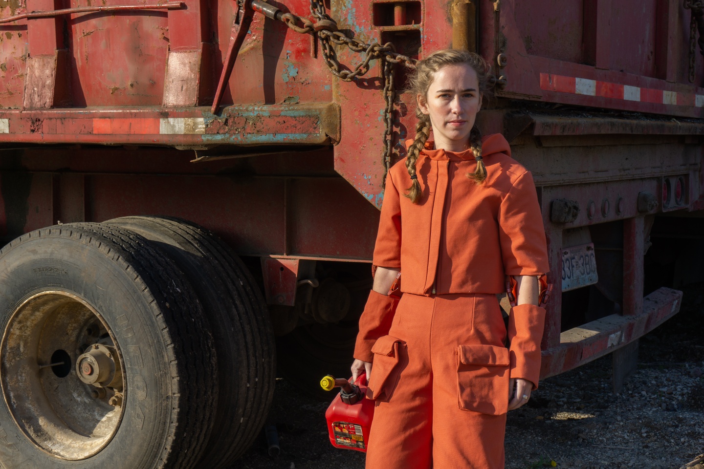 Model wears an a construction orange outfit consisting of pants with cargo pockets and a hooded cropped jacket with zipper and detachable sleeves below the elbow. Model stands in front of a dump truck clutching a gas can.