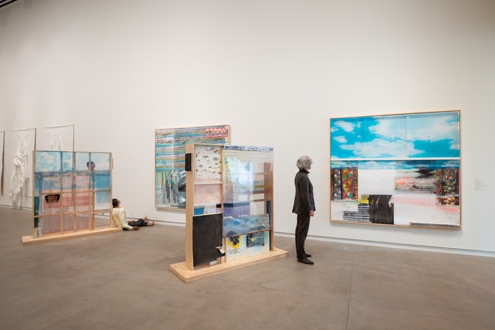 A person inspects an installation made of two free-standing wooden shelves and two flat works on the wall. All are constructed of a variety of materials, including wood, papier mache, plastic film, wallpaper, and paint.