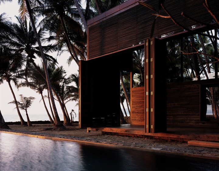 Exterior view of a house with a wooden louvered structure anchored to a stone platform. Multiple palm trees can be viewed to the left of the house.