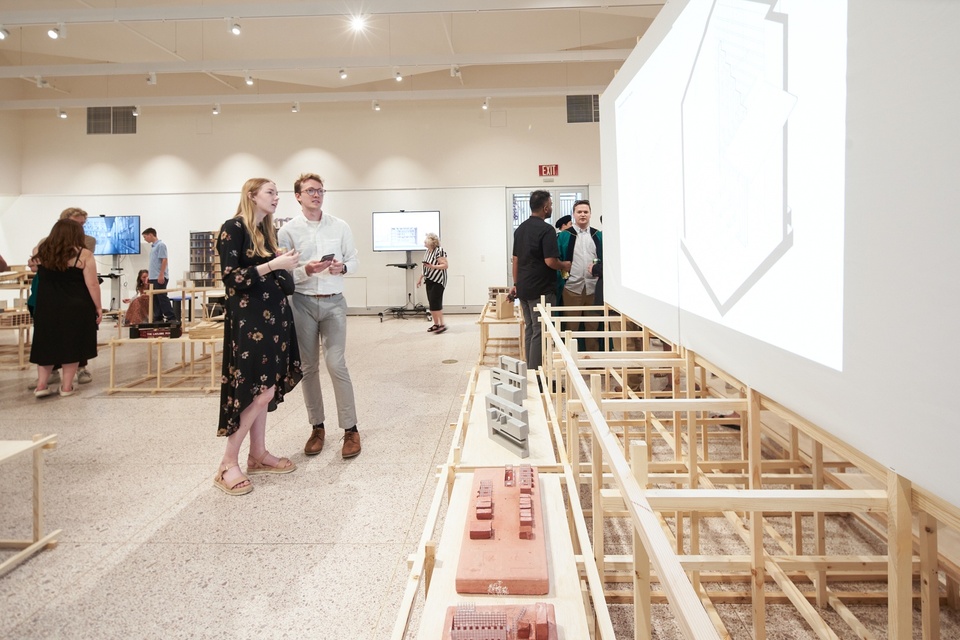 Two people stand together and view an architectural drawing projected onto a freestanding screen in the middle of a gallery space. Small physical models are placed along a shelf in front of the screen.