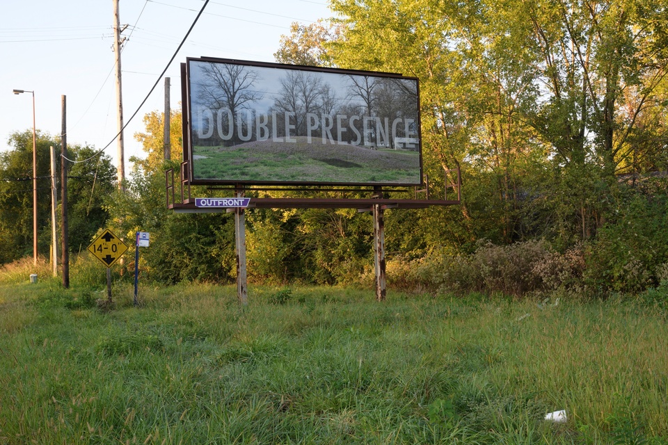 A picture shows a billboard with a picture of a mound and the words "Double Presence" across the image. Around the billboard is grass and overgrown bushes. There is a bus stop and a bridge height sign to the left of the billboard.