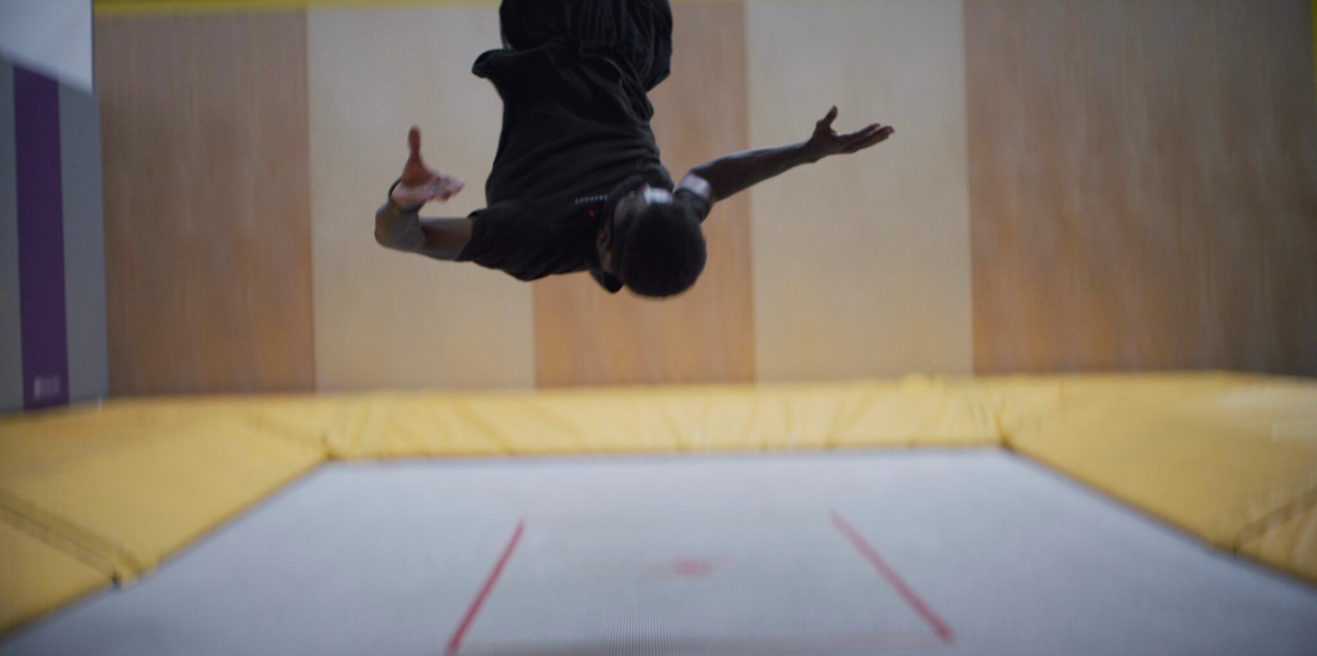 Film still of a person in black suspended upside down from a ceiling above a white and yellow space on the ground.