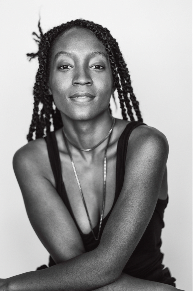 A portrait of actor Krysten Peck, a black woman sitting with her arms crossed low across her lap. She wears a black tank top and long necklace. She smiles slightly with eyebrows raised.