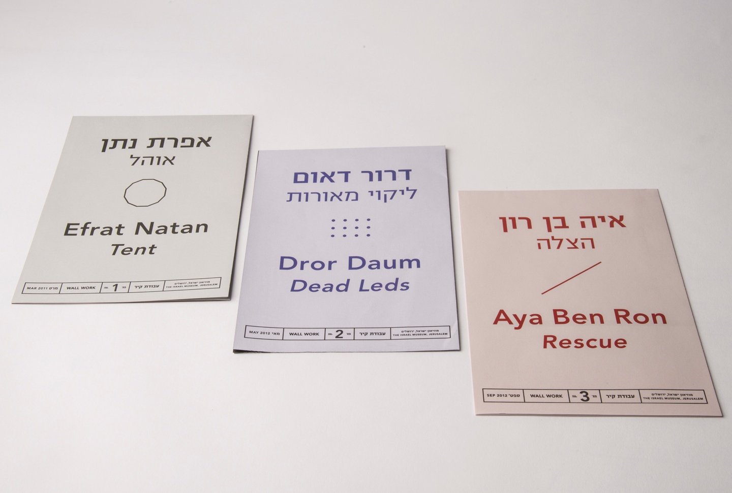 Three pastel-toned booklets laid out on a white background (from left to right, in gray, lavender/blue, and orange/coral/red). The covers include artists' names with titles below, in both Hebrew and English, a geometric pattern in the center, and supplementary information at the bottom in a row.