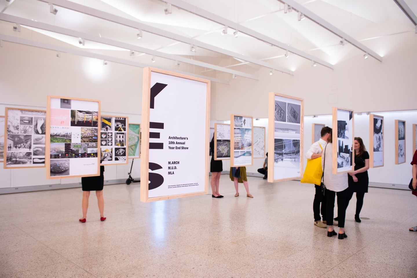 An exhibition of architectural drawings. Work hangs in simple, blonde wood frames, suspended from the pleated ceiling by invisible lines. People are gathered in small groups around the work.