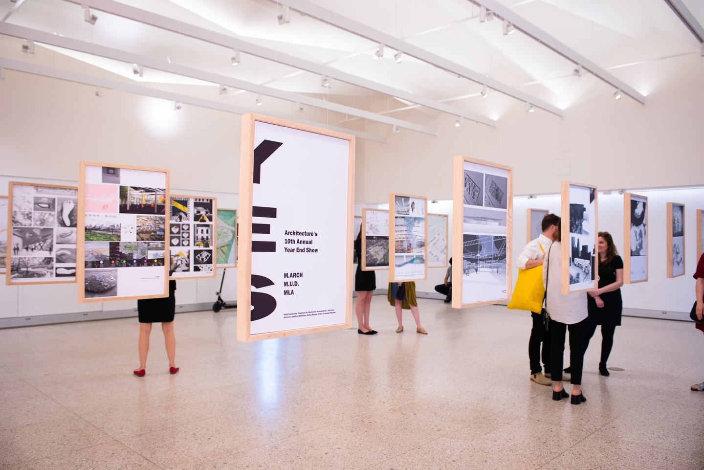 An exhibition of architectural drawings. Work hangs in simple, blonde wood frames, suspended from the pleated ceiling by invisible lines. People are gathered in small groups around the work. 