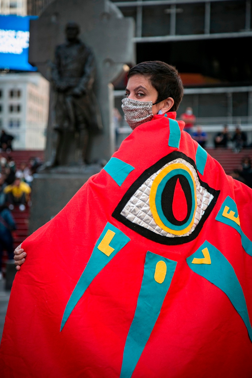 A photograph of a person wearing a mask and a red cape with an eye and “LOVE” in yellow. Behind them, out of focus, stands a tall statue.