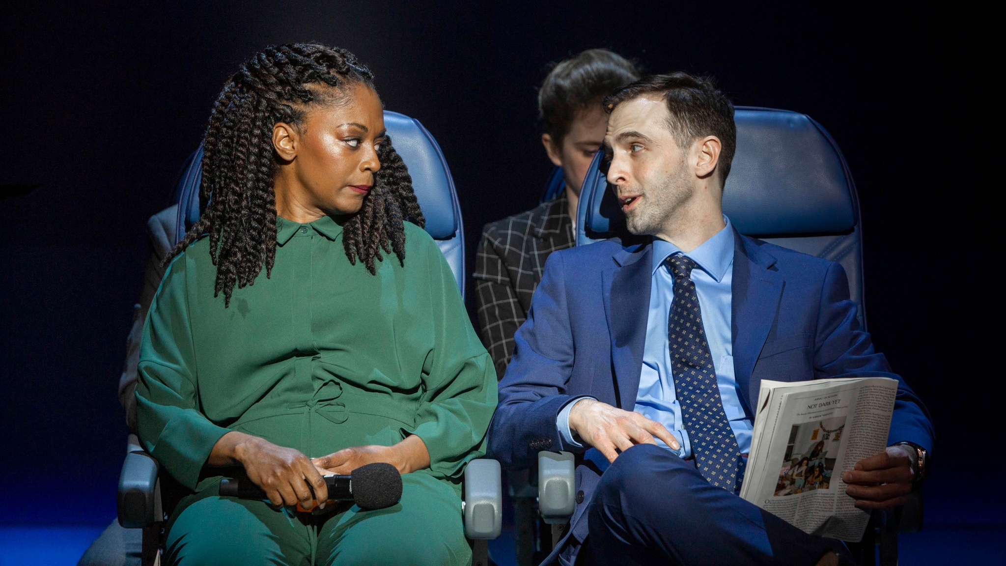 A Black woman in a green jumpsuit and a white man in a blue suit turn toward each other while sitting in airplane chairs. They are illuminated by a spotlight. 