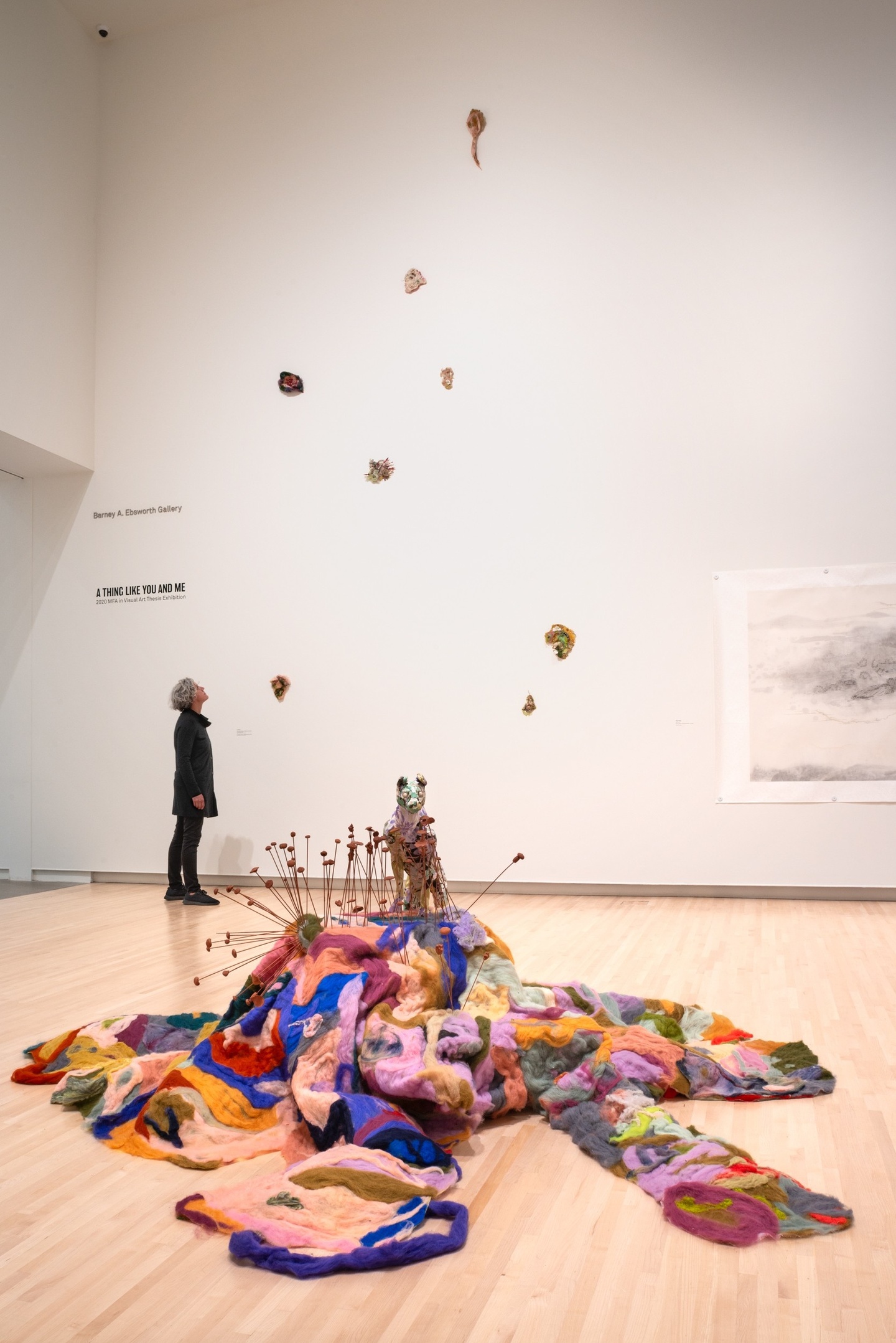 A large multi-colored felt sculpture on the gallery floor. Wires poke out of it like mushrooms and a multi-colored animal perches above it
