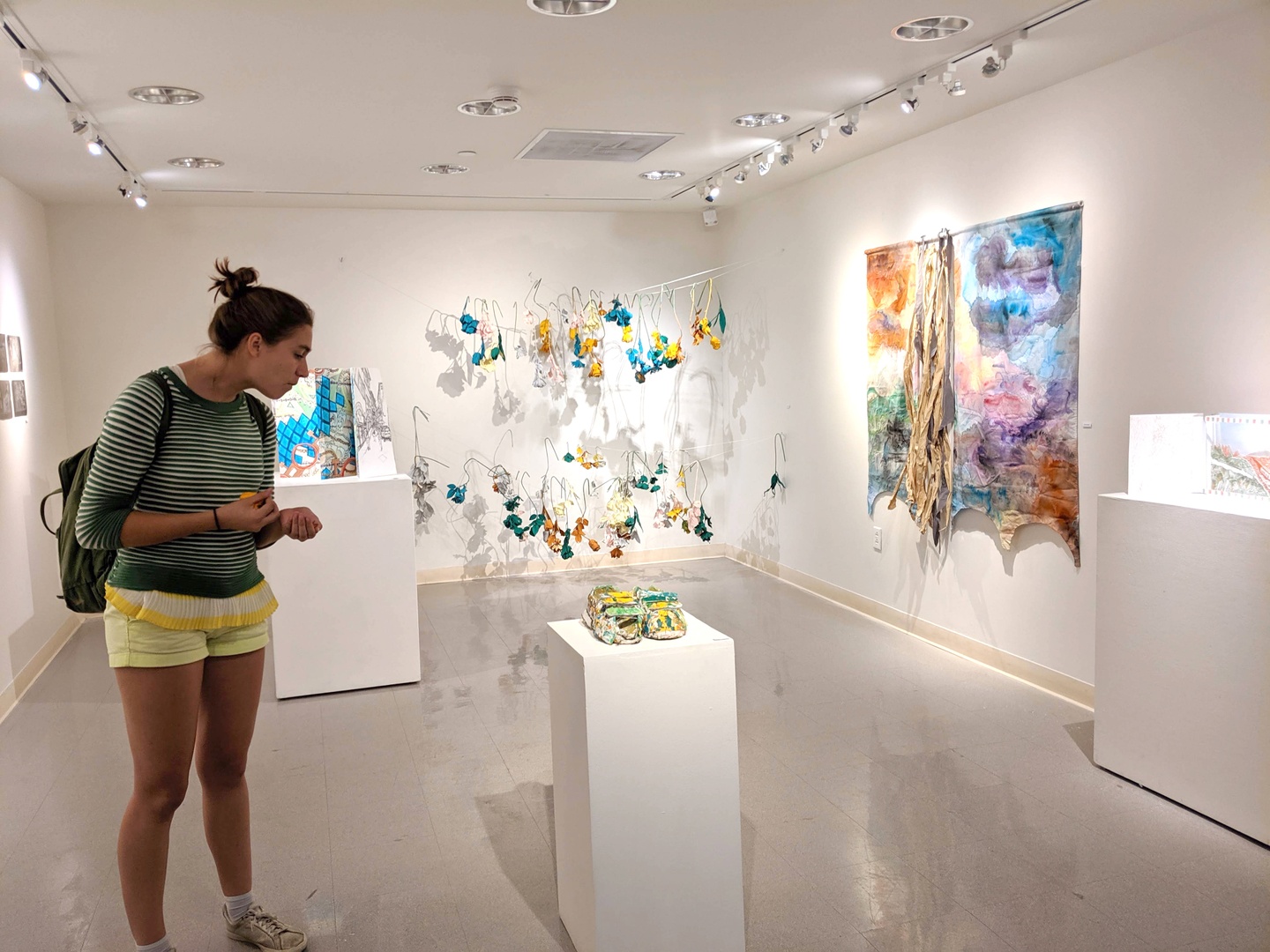 A person looks at an object on a white plinth in a small white-walled gallery space.