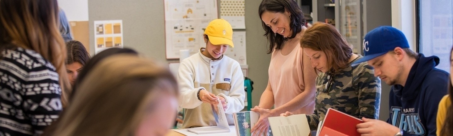 Students in a classroom leafing through design books laid out on a white sheet of paper on a wooden table.