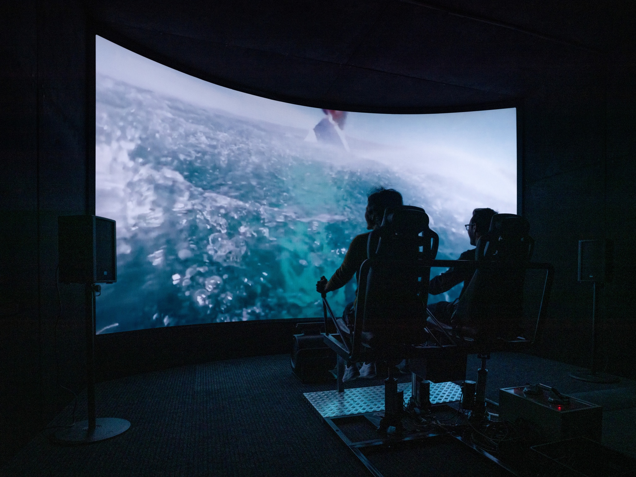 Twp people seated in a flight simulator in front of a large screen showing close-up video of ocean water