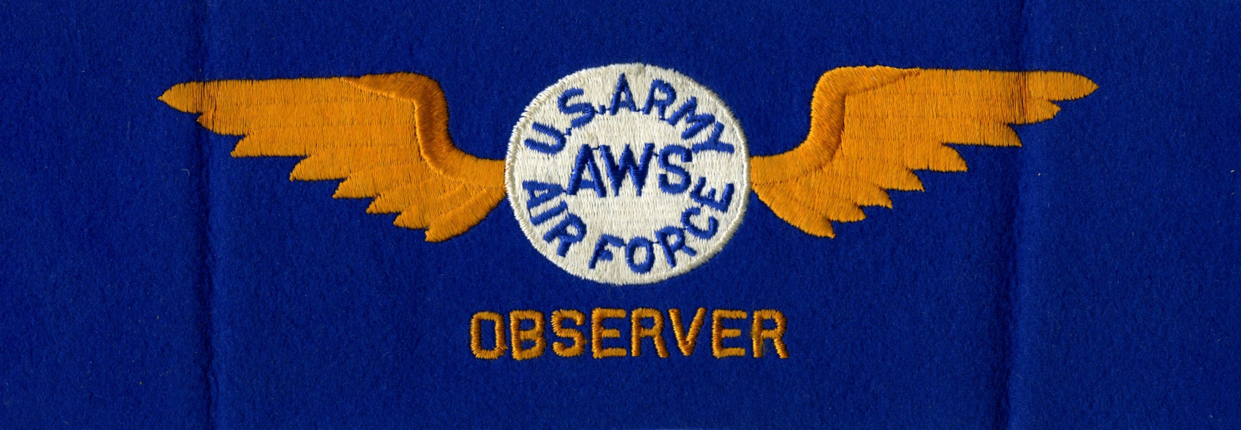 An embroidered white circle on blue fabric has the words “U.S. ARMY AWS AIR FORCE” embroidered in blue letters. A pair of pale orange wings are embroidered on either side of the circle, and below it says “OBSERVER” in the same orange thread.