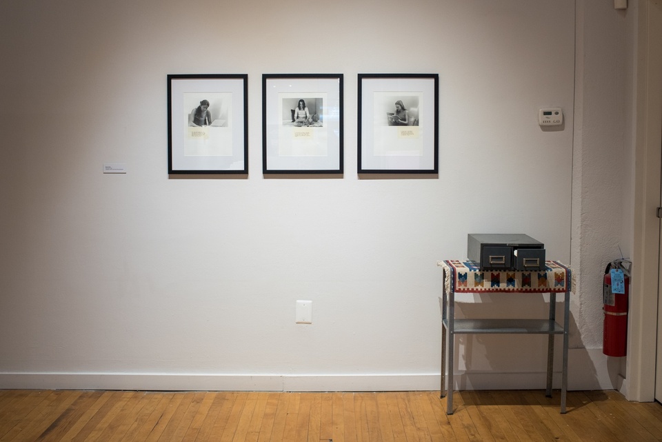 Three small black and white photographs of women sitting at desks hang on a white wall. Beside them is a small metal shelf with a 2-drawer card catalog placed upon it.