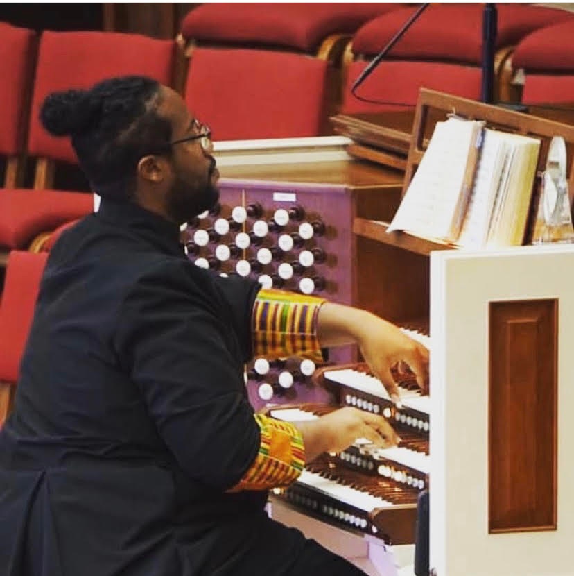 A Black man sits at an organ with is back to the camera while he plays the instrument. 