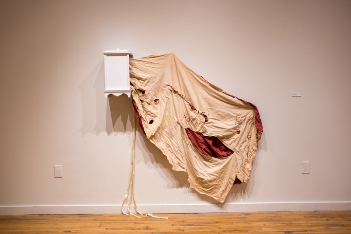 A small wooden cabinet is affixed to the wall and a large, tattered piece of beige and red fabric is draped out of it.