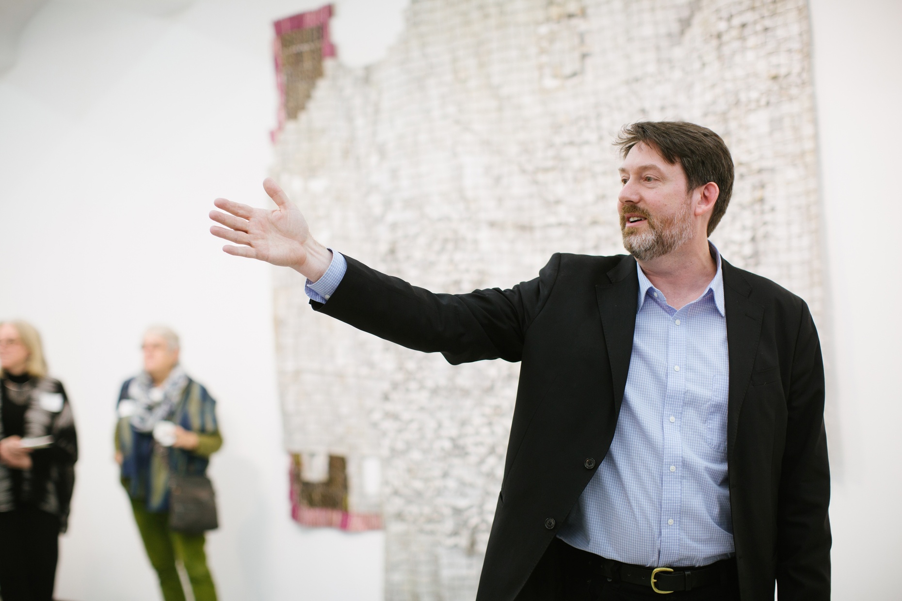 A middle-aged, white man stands with one arm raised in front of a large artwork hung on a white wall.