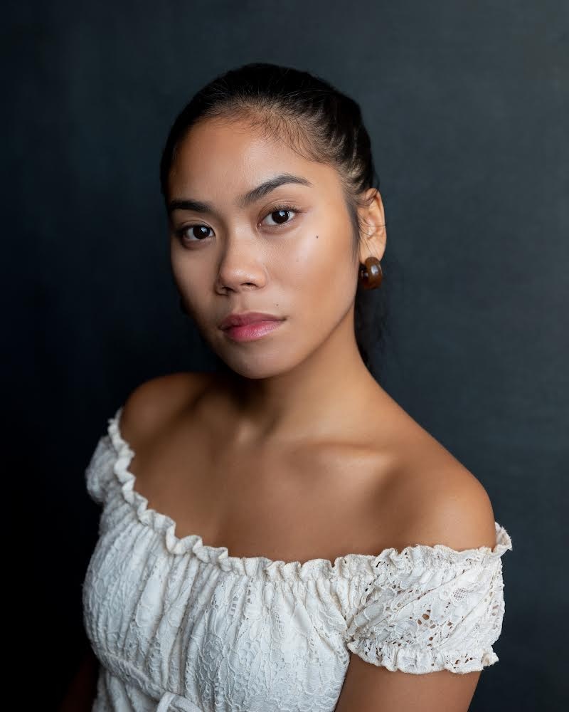 A headshot of actor Melanie-Joyce Bermudez. She turns slightly toward us with one shoulder and raises an eyebrow quizzically. She wears a frilly blouse that shows her bare shoulders. 