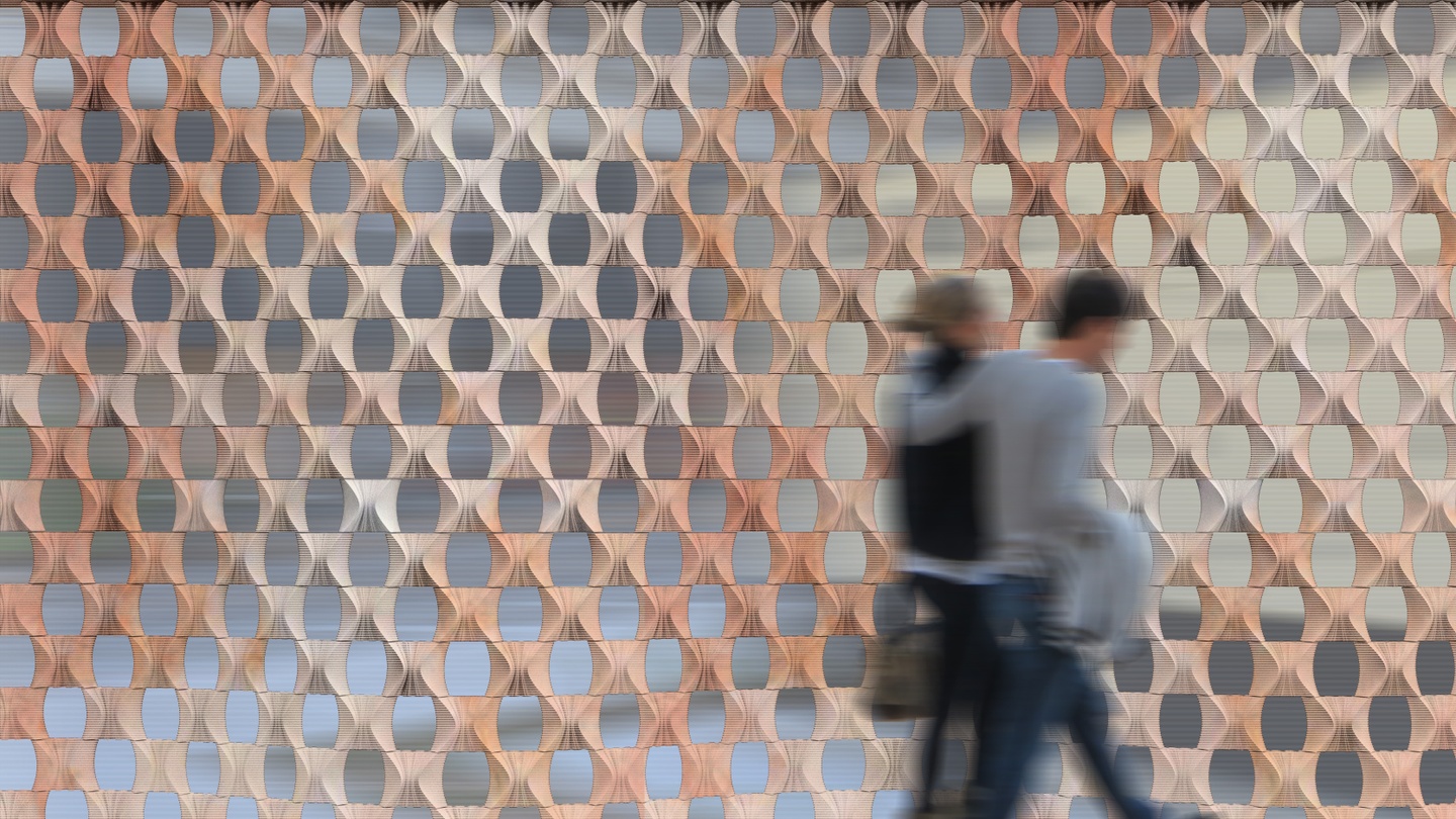 Digital rendering of two people walking in front of a 3D printed masonry screen wall, with a honeycomb like pattern with openings.